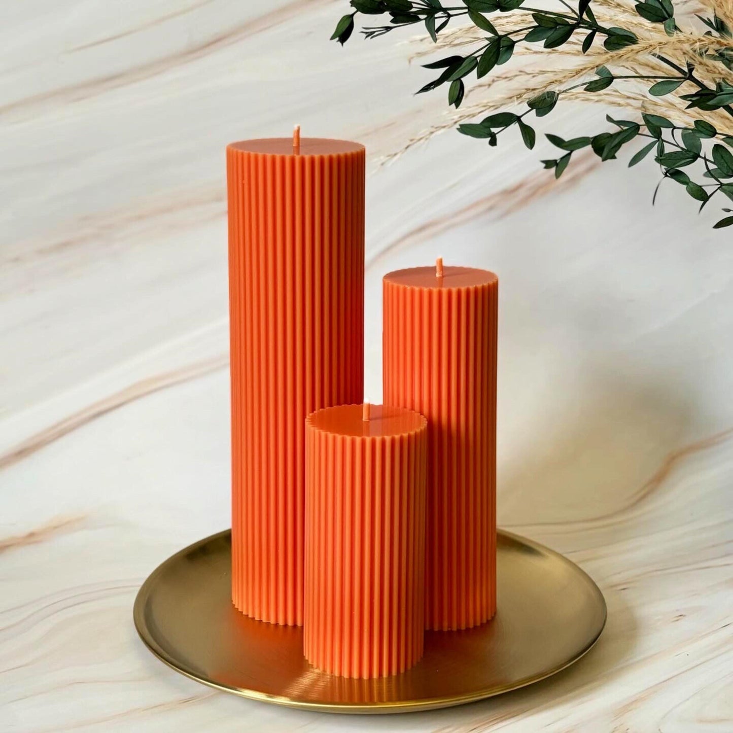 Three tall orange candles with a textured surface are displayed on a gold plate, with a plant branch visible in the background.