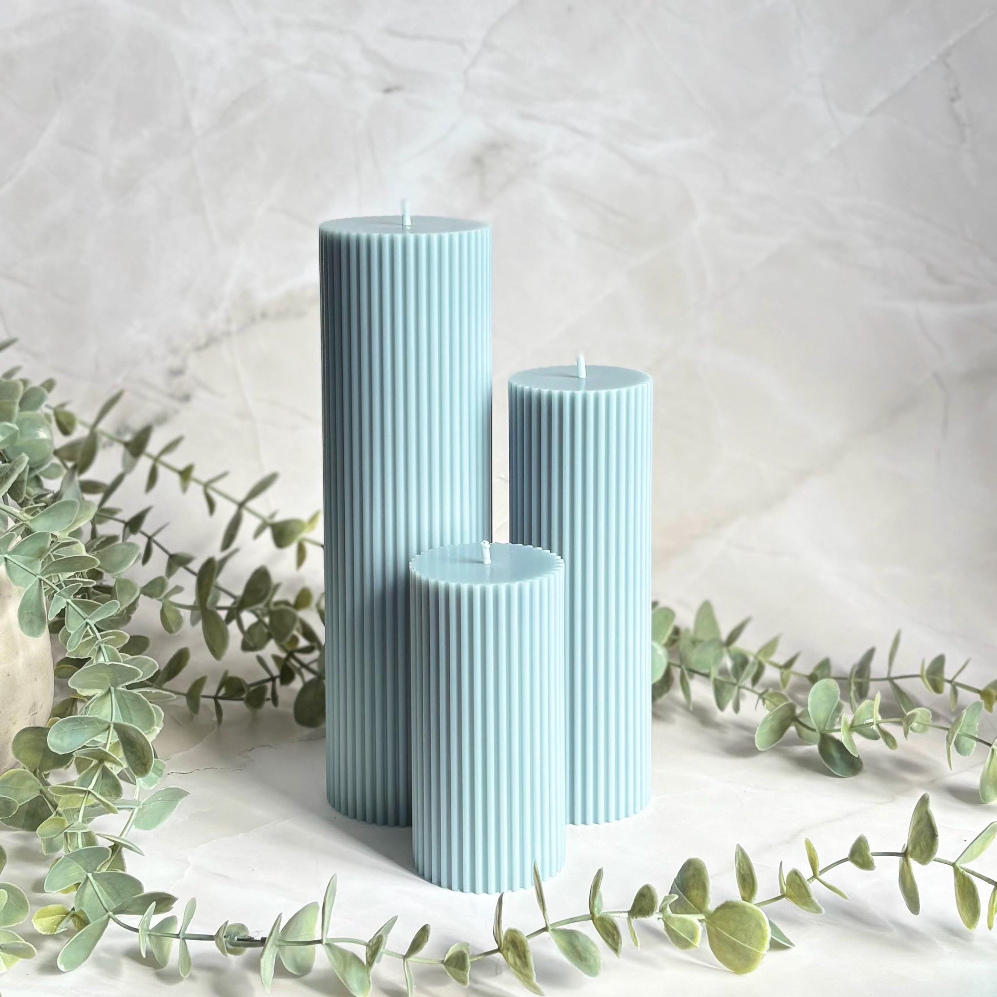Three cylindrical candles in a light blue color are placed on a white surface, surrounded by greenery and a white marble background.
