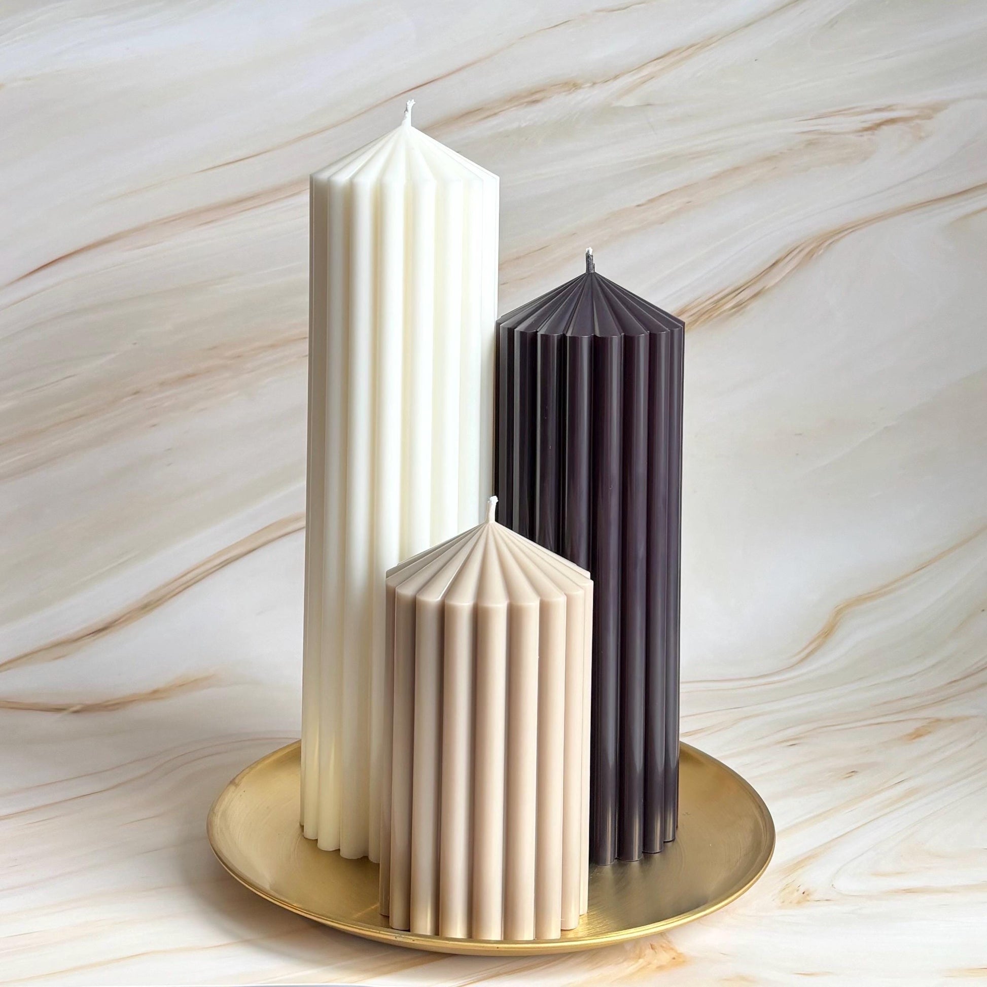 Three candles of different colors - white, beige, and dark - are arranged on a gold plate against a marble background.
