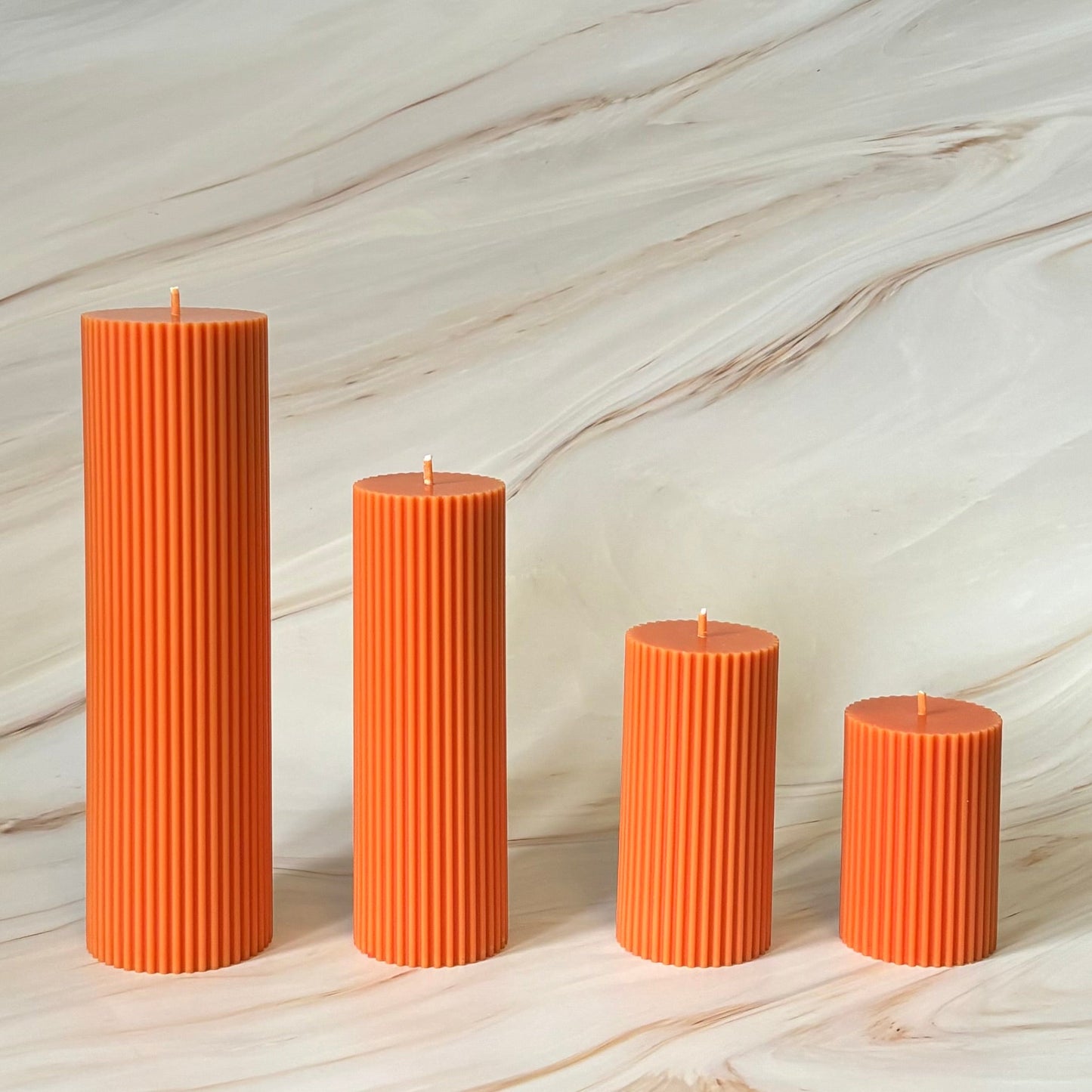 Four orange pillar candles of varying sizes are arranged in a row against a marble background.