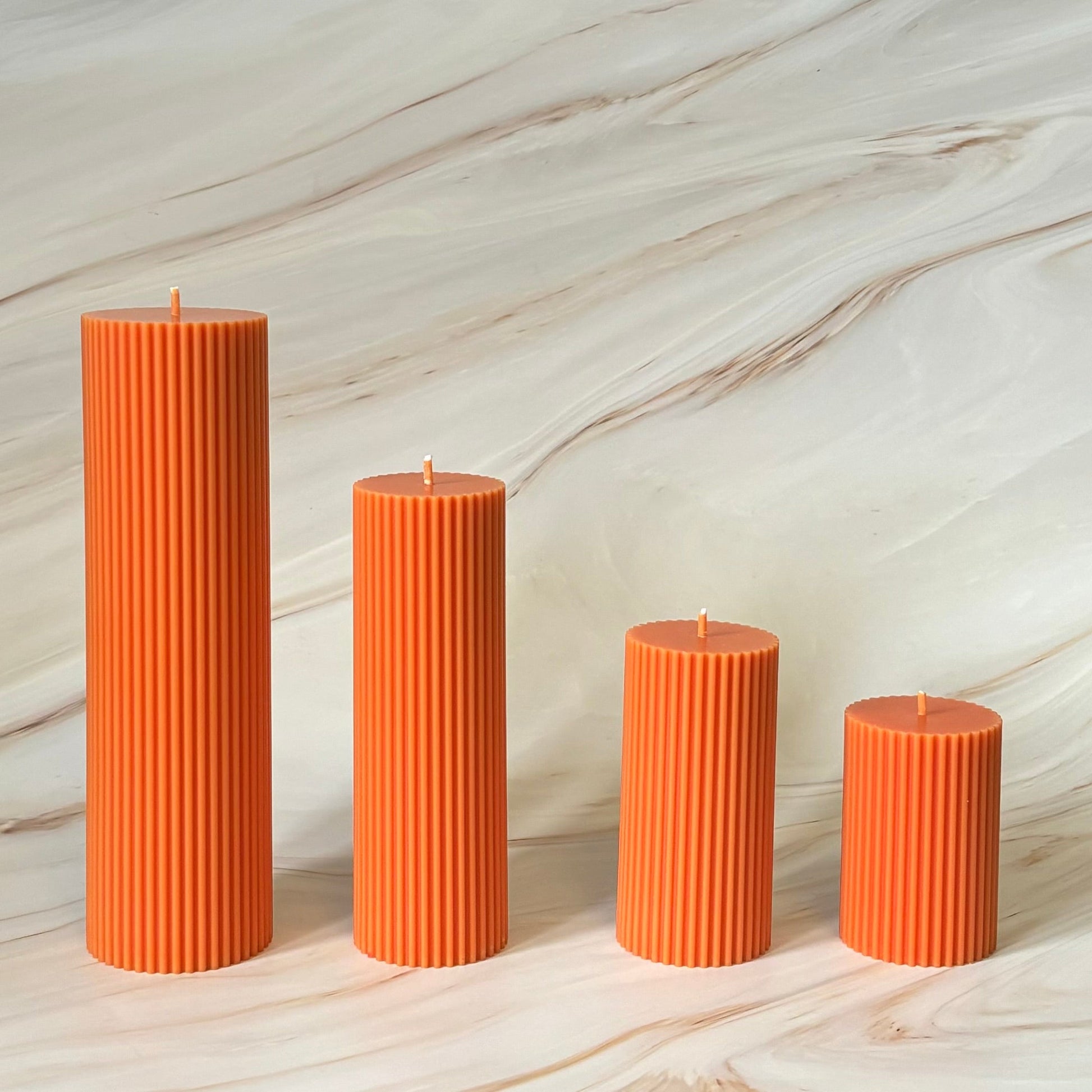 Four orange pillar candles of varying sizes are arranged in a row against a marble background.