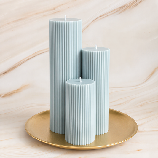 Three tall, cylindrical candles in a light blue color are placed on a gold plate against a marble background.