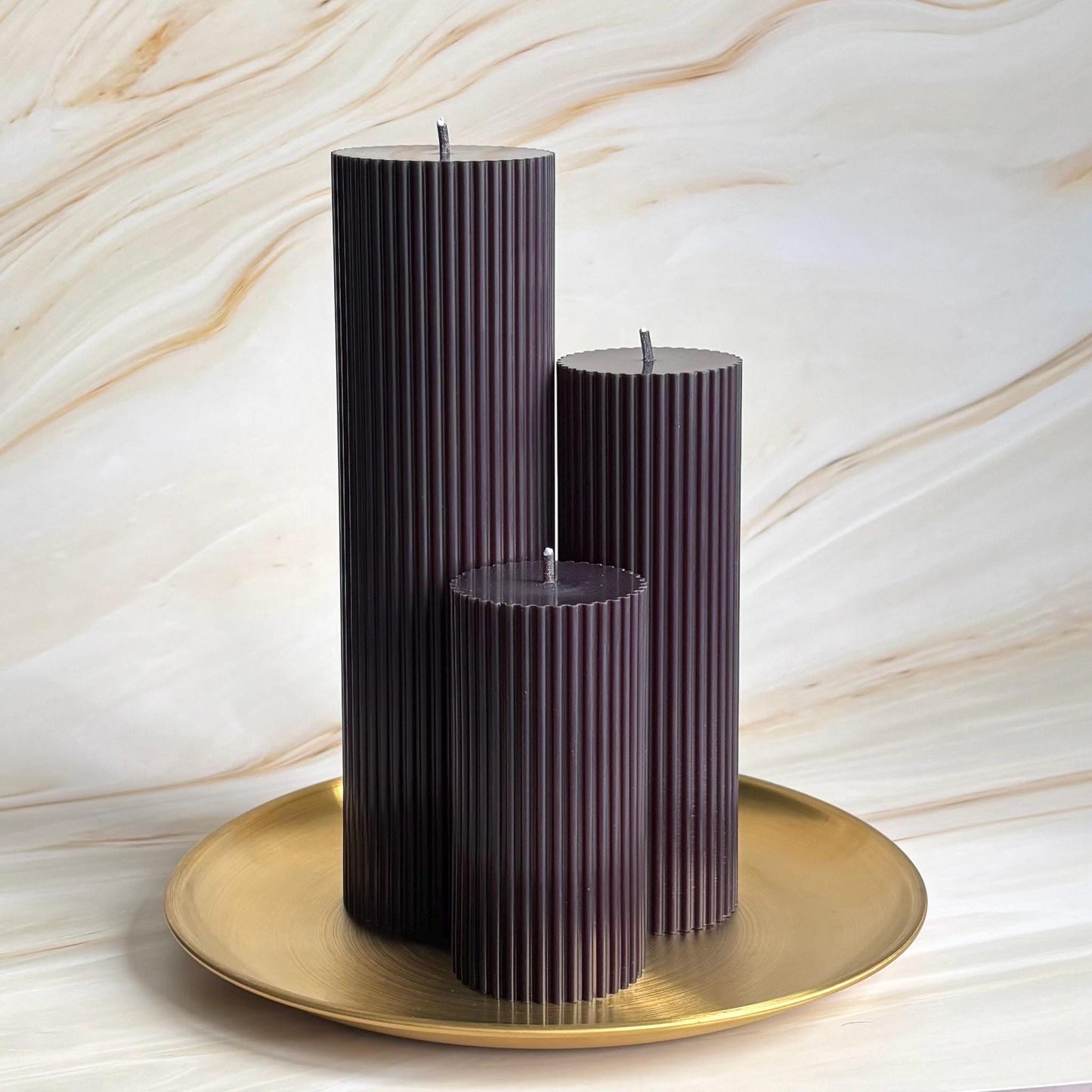 Three tall, cylindrical candles in a dark brown color are displayed on a gold plate against a marble background.