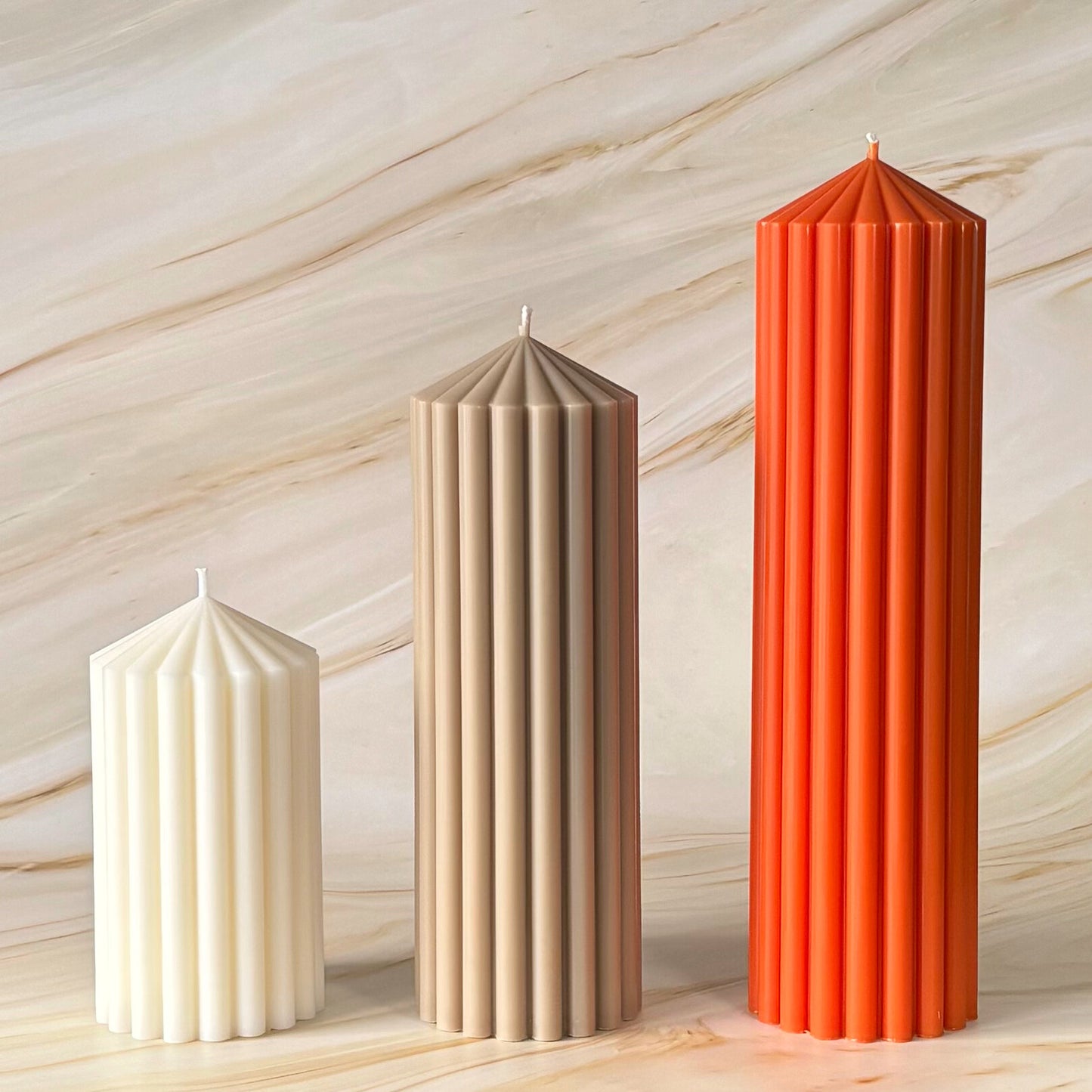 Three tall candles of different colors - white, beige, and orange - stand upright against a marble background.