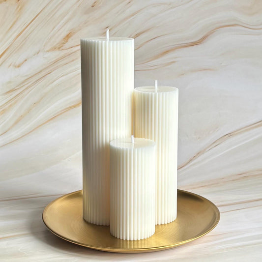 Three tall, cylindrical white candles are displayed on a gold plate against a marble background.