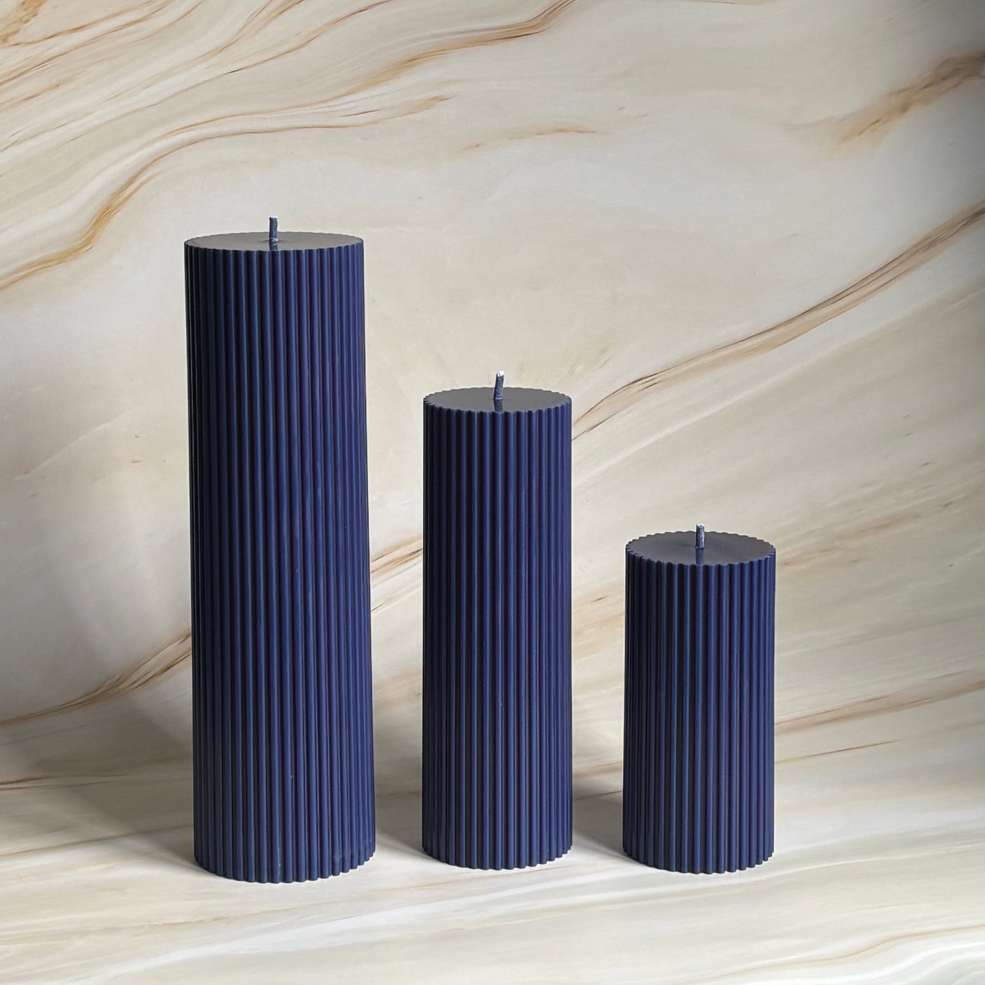 Three tall, cylindrical candles in a dark blue color are arranged in a row against a marble-like background.