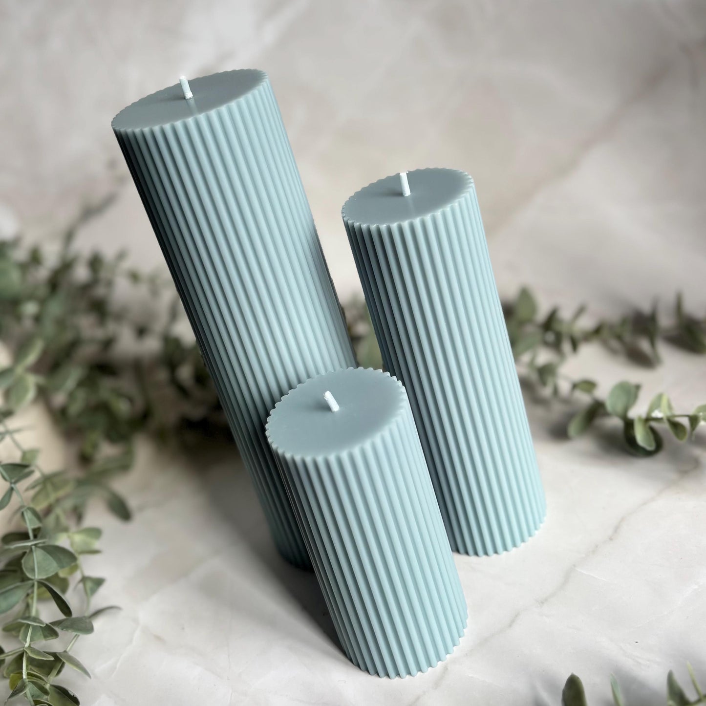 Three cylindrical candles in a light blue color, arranged on a white surface with some greenery in the background.