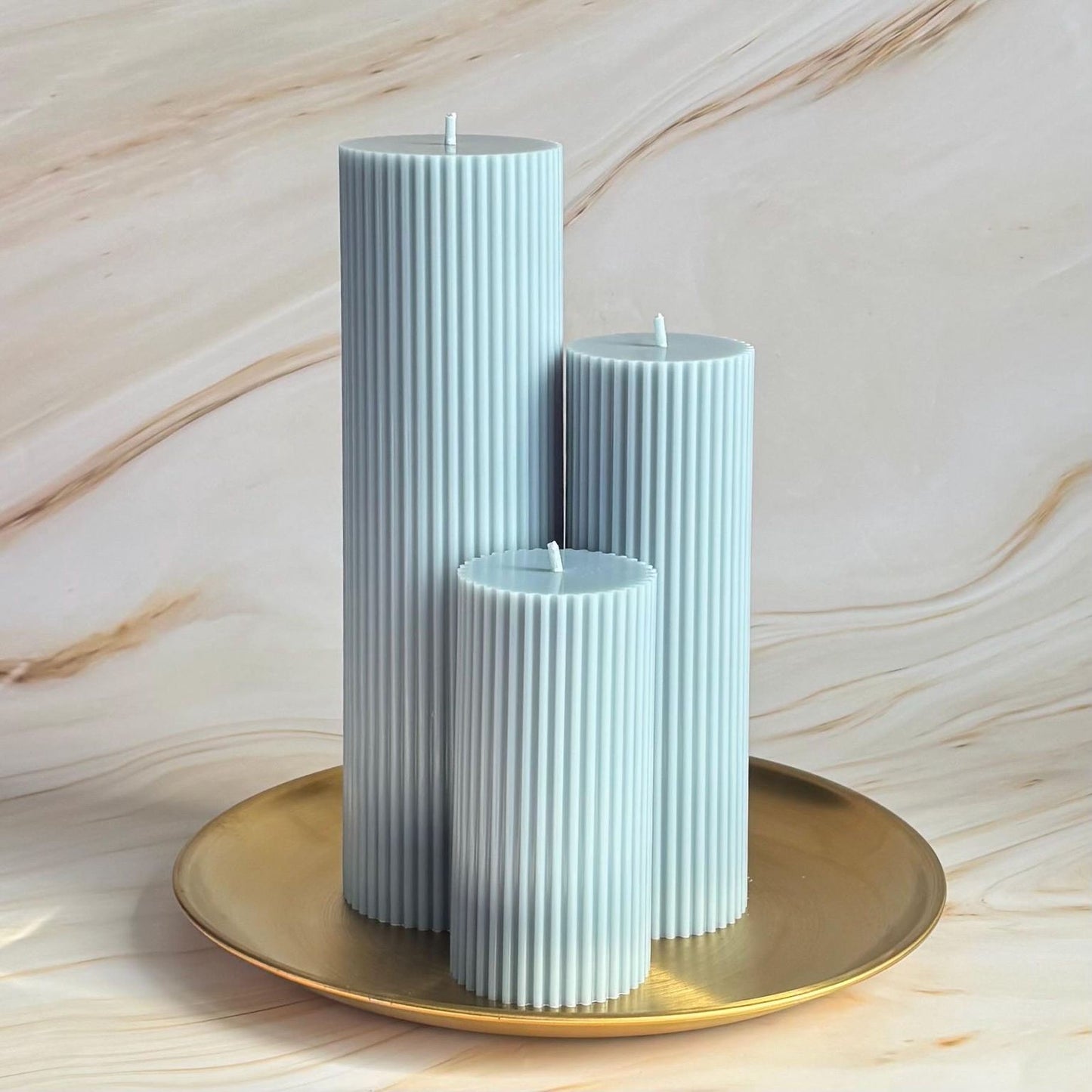 Three tall, cylindrical candles in a light blue color are displayed on a gold plate against a marble background.