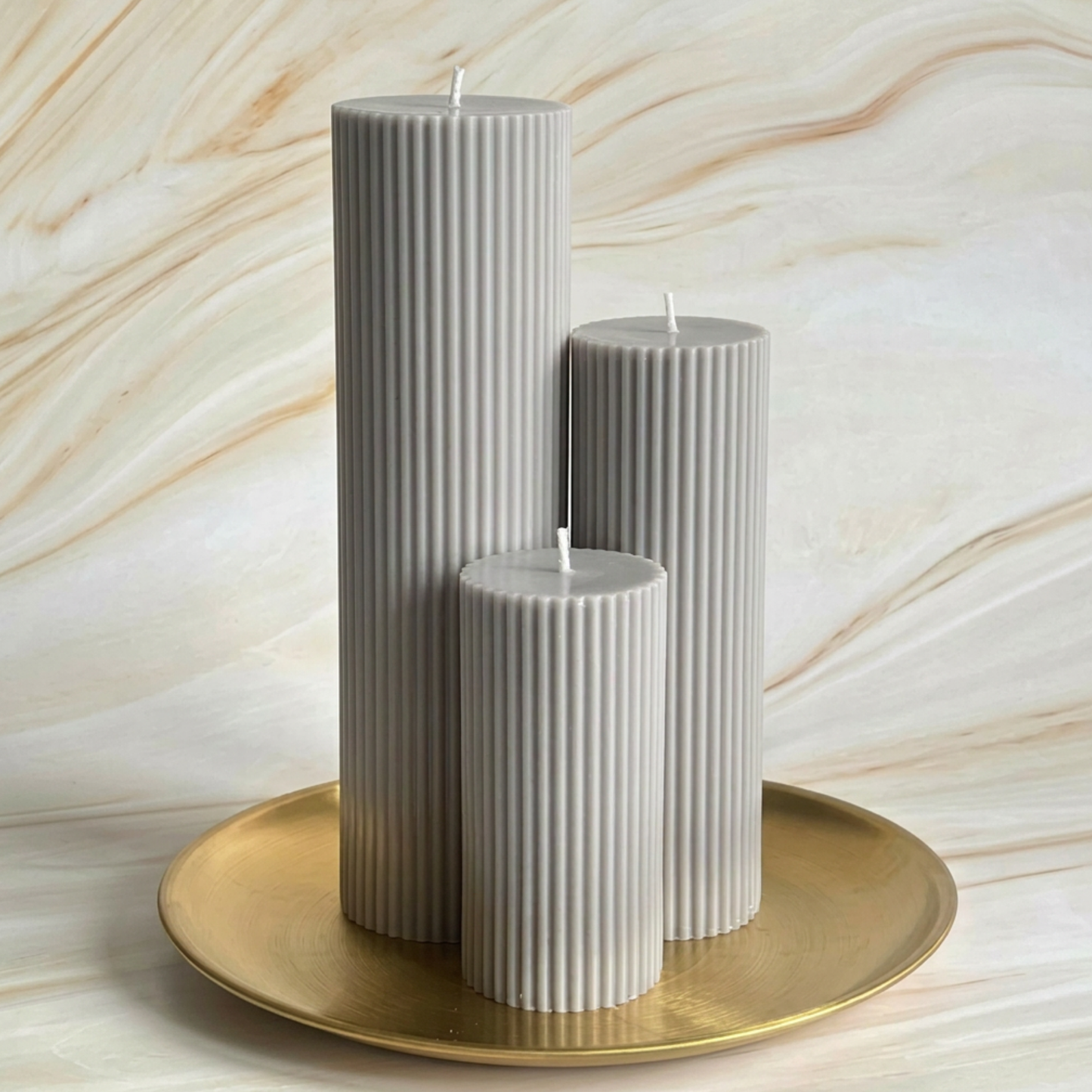 Three tall, cylindrical candles with a gray color are placed on a gold plate against a marble background.