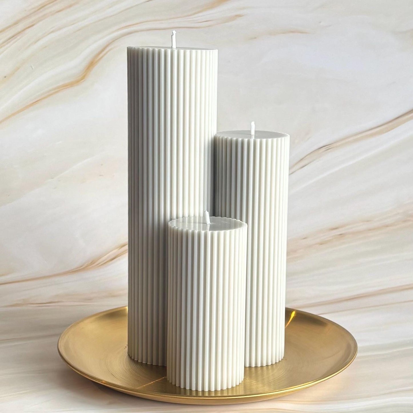 Three white cylindrical candles with a striped design are placed on a gold plate against a marble background.