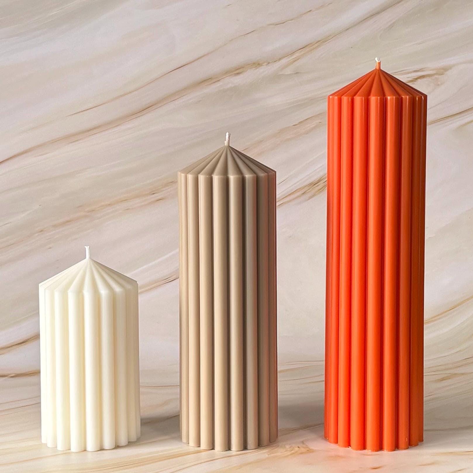 Three tall candles with different colors - white, beige, and orange - stand upright against a marble background.