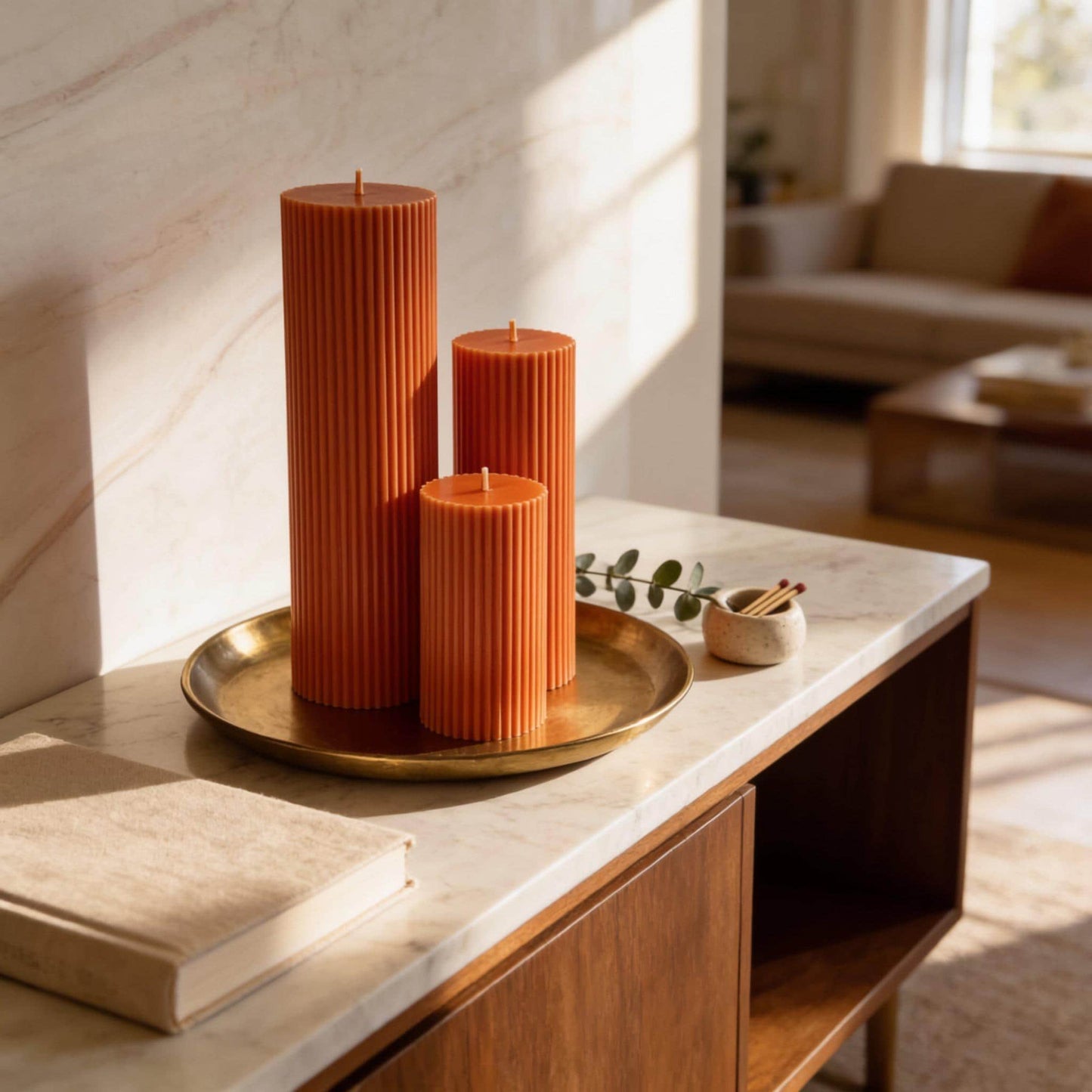 a marble countertop with three orange cylindrical candles and a small white bowl containing a plant. The candles are arranged on a gold tray, which is placed on a wooden cabinet. The background shows a living room with a couch and a window.
