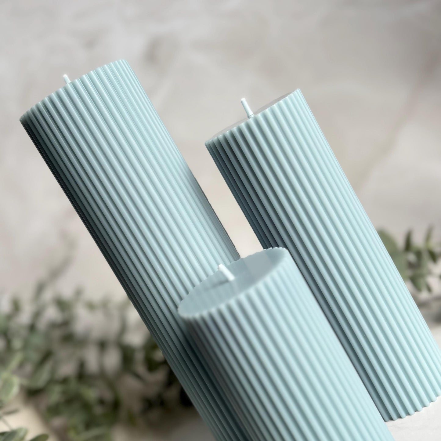 Three cylindrical candles with a light blue color are placed together, with a blurred background that includes some greenery.