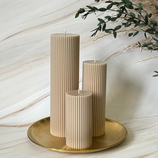 Three beige pillar candles with a cylindrical shape and vertical lines are displayed on a gold plate, with a plant in the background.