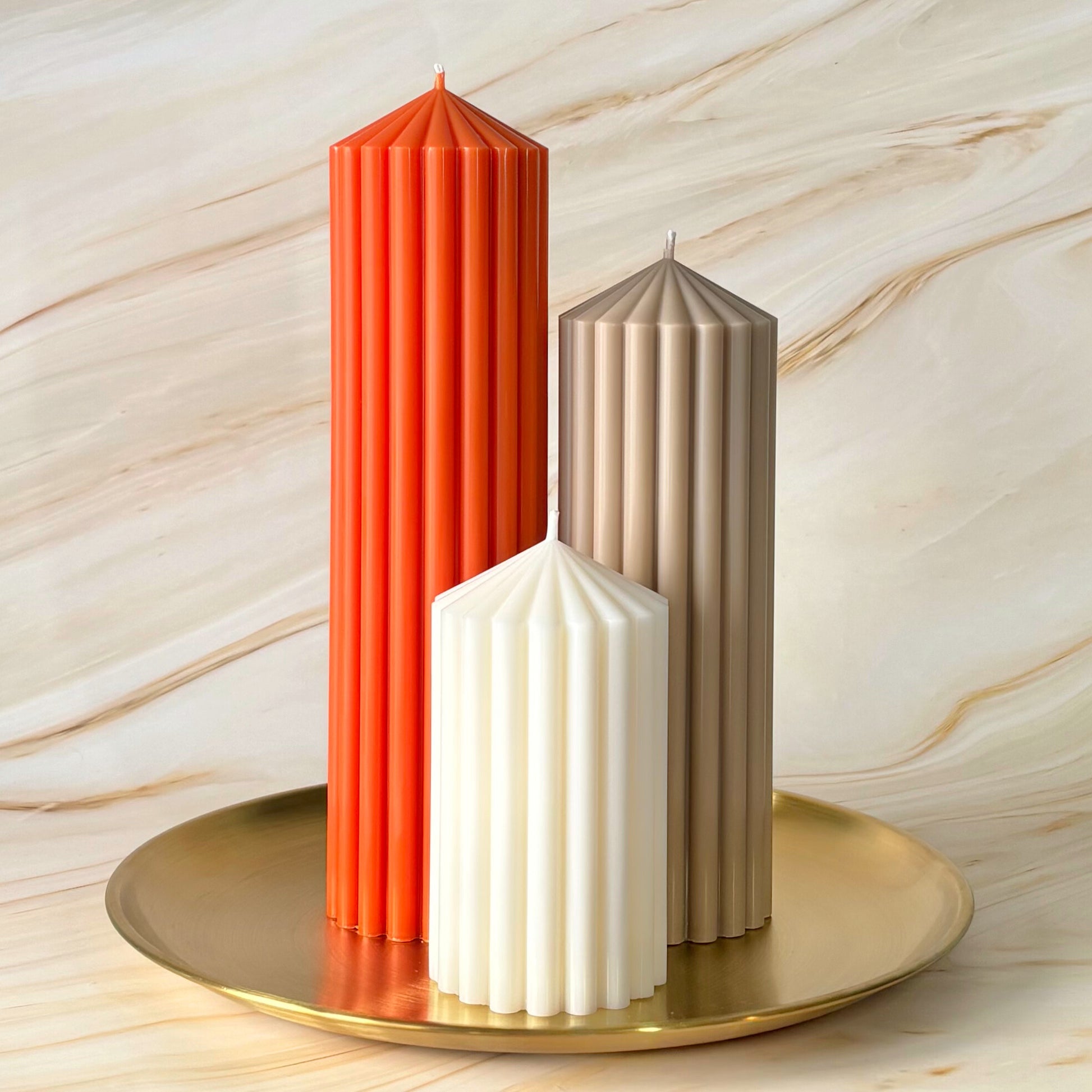 Three tall candles in different colors - red, beige, and white - are arranged on a gold plate against a marble background.