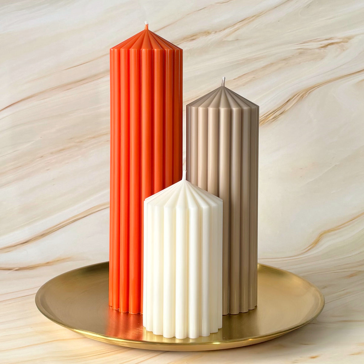 Three tall candles in different colors - red, beige, and white - are arranged on a gold plate against a marble background.