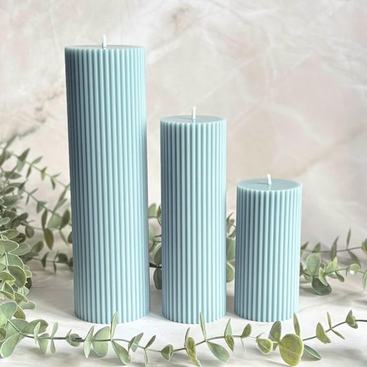 Three cylindrical candles in a light blue color, placed on a white surface with greenery in the background.