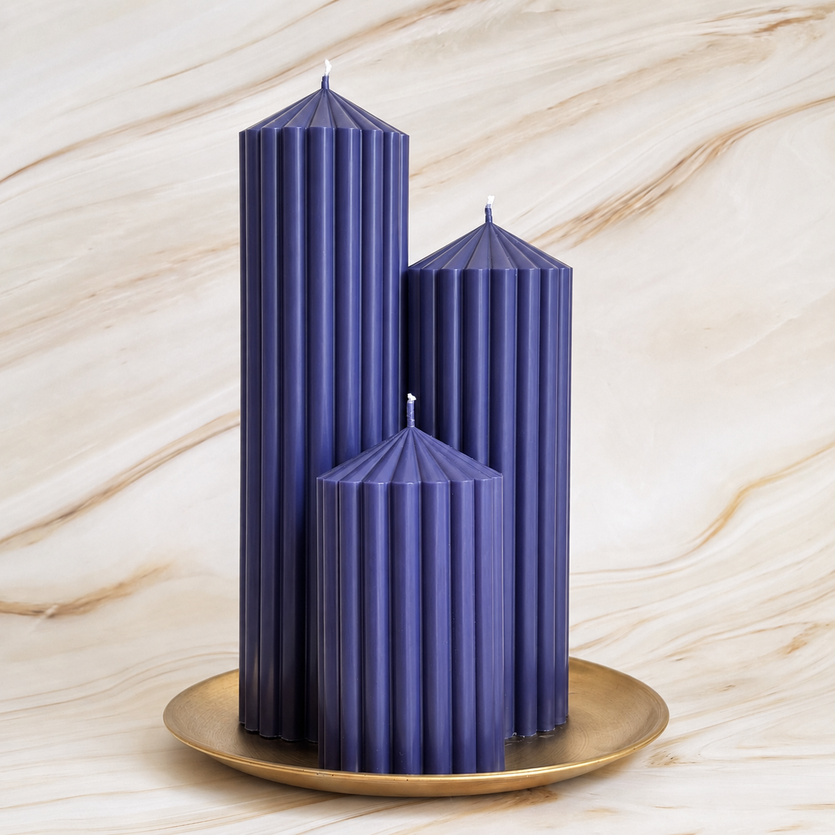 Three tall, cylindrical candles in a dark blue color are displayed on a gold plate against a marble background.