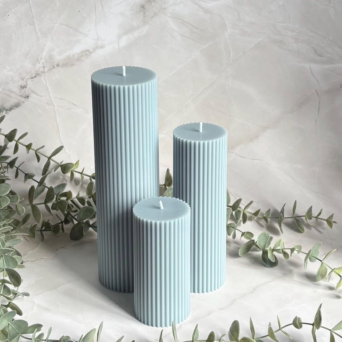Three cylindrical candles in a light blue color are placed on a marble surface, surrounded by greenery.