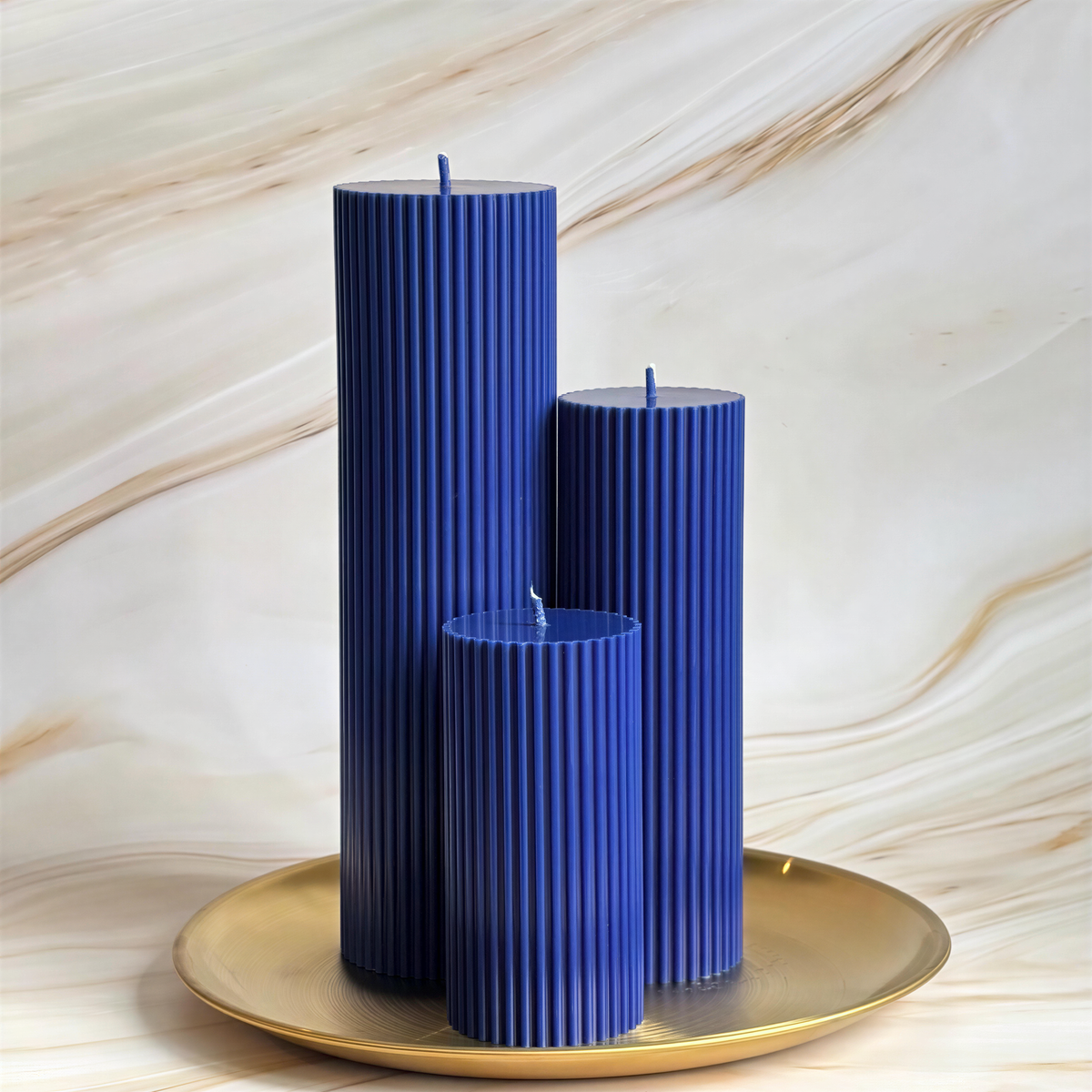 Three tall, cylindrical blue candles are displayed on a gold plate against a marble background.