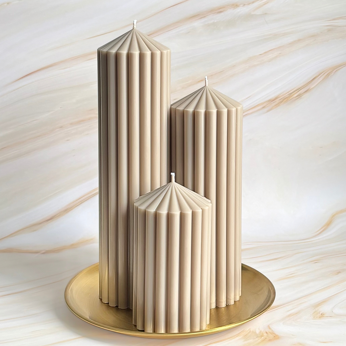 Three tall, cylindrical candles with a beige color are displayed on a gold plate against a marble background.