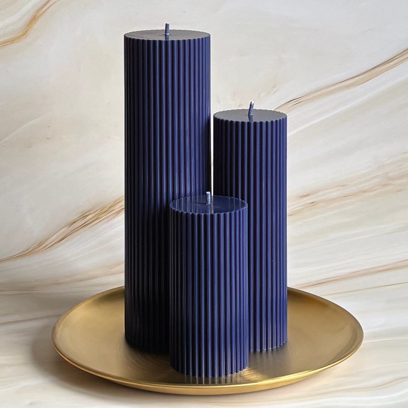 Three tall, cylindrical candles in a dark blue color, placed on a gold plate against a marble background.