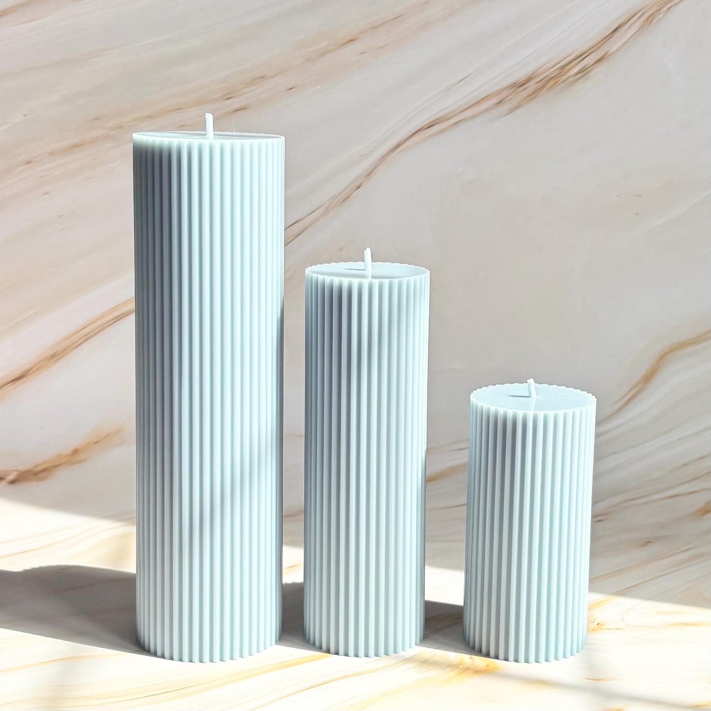 Three cylindrical candles with a light blue color are arranged in a row against a marble background.