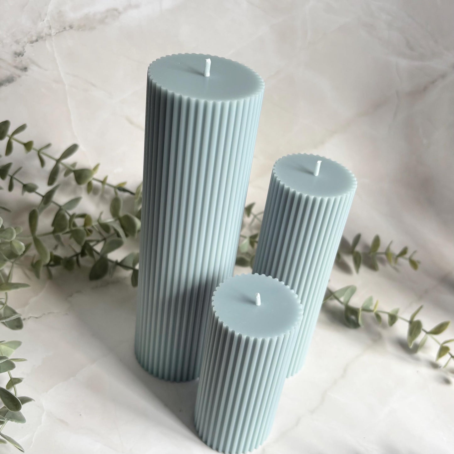 Three cylindrical candles in a light blue color, placed on a white marble surface with some greenery in the background.